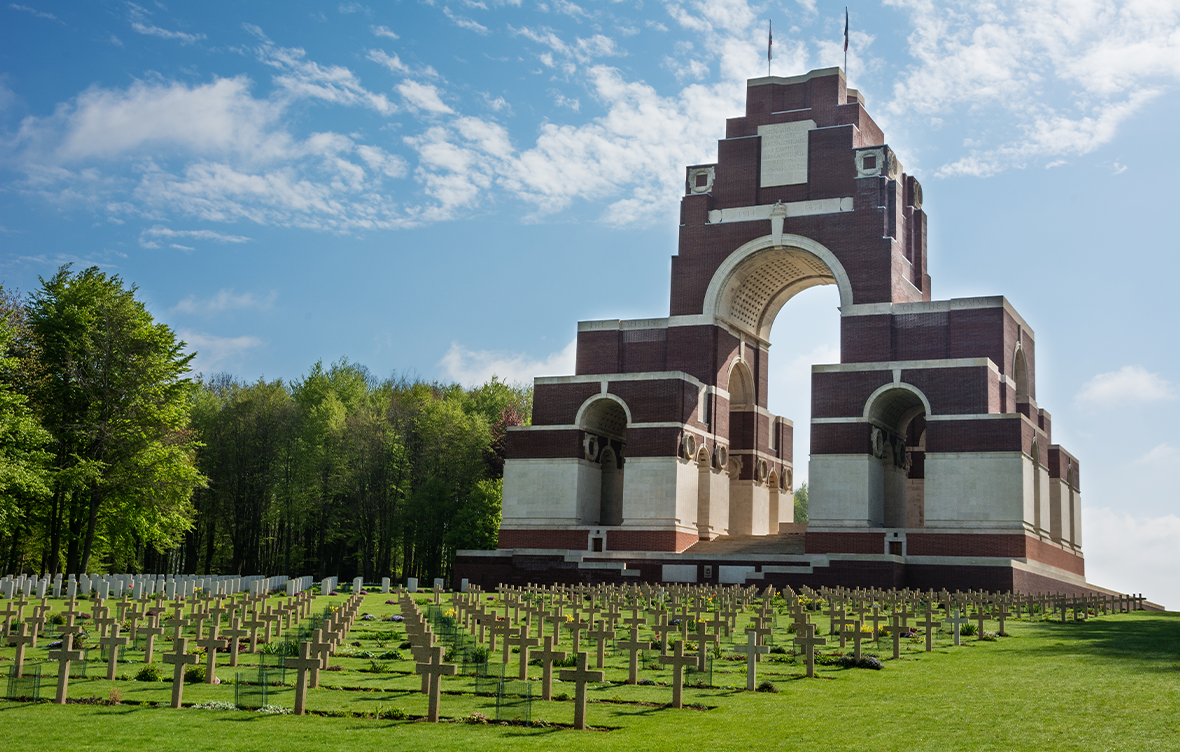 Large arch made of red and white brick with graves marked with wooden crosses lying across a field below